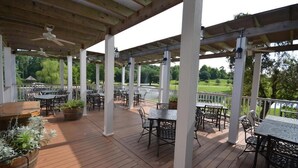Eine überdachte Terrasse im Freien mit Holzboden, Metalltischen und -stühlen, umgeben von weißen Säulen und einem Geländer.