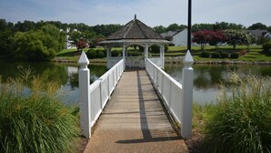 Eine Holzbrücke führt zu einem Pavillon über einem stillen See, umgeben von Grünflächen und Häusern.