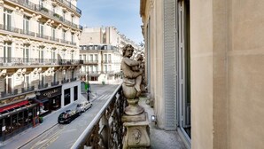 Un balcó amb una estàtua de querubí, amb vista a un carrer ple d'edificis i botigues.