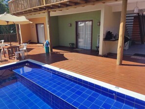 A pool area with a wooden deck, a covered seating area, and a door leading to an interior space. - Sítio de Praia (Porto Seguro)