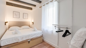 Een moderne slaapkamer met een houten bedframe, een hoofdeinde, een nachtkastje met een lamp, een raam met dunne gordijnen en een wandplank met kledinghangers.