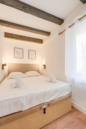 A bedroom with a wooden headboard, two framed pictures on the wall, a bed with white linens, and a window with sheer curtains.
