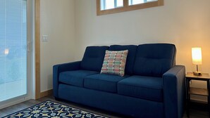 A living room with a blue sofa, a patterned throw pillow, a small table with a lamp, and a window above the sofa.