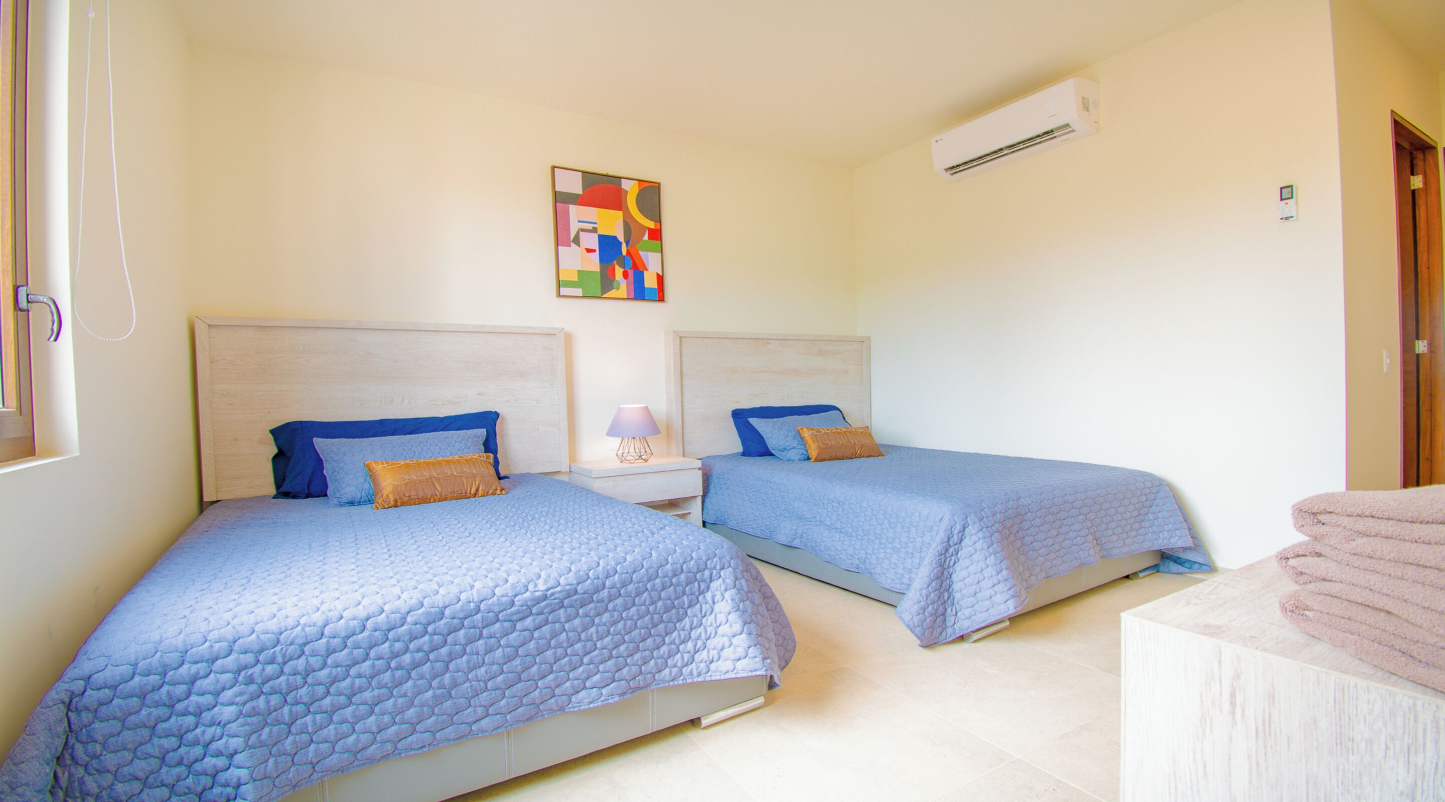 Un dormitorio con dos camas, cada una con colcha y almohadas azules acolchadas, una mesita de noche con lámpara, y un cuadro abstracto de colores en la pared.