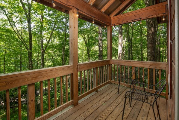 A wooden deck with metal chairs and a railing, surrounded by a forest. - Red Feather Cabins 199 (Boone)