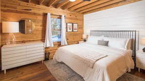 A bedroom with a wooden ceiling, a bed with white bedding, a nightstand with a lamp, a dresser, and a window with curtains.