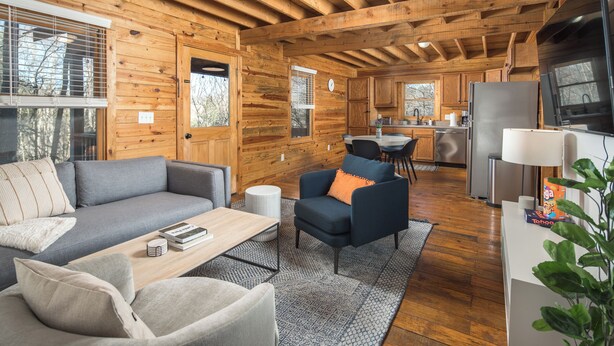 A cozy cabin interior with a living area featuring a sofa, armchair, and coffee table, a kitchen with modern appliances, and a dining area.