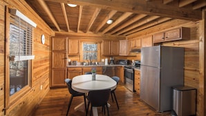 A wooden cabin kitchen with a round dining table, built-in cabinets, a refrigerator, and a window with blinds.