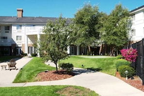 A courtyard with a lawn, a tree, a bench, and a building with a balcony.