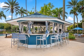A covered outdoor bar area with seating, surrounded by palm trees and a pool.