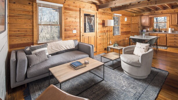 A cozy cabin interior with a sofa, armchair, coffee table, and a kitchen area with wooden cabinets and a window.