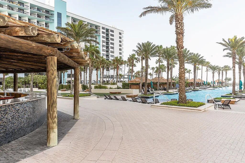 A resort pool area with palm trees, lounge chairs, and a modern building in the background.