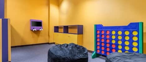 Una habitación con una pared amarilla, un mueble de televisión púrpura, un tablero de juego azul y amarillo, y dos puffs negros.