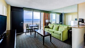 Ein Hotelzimmer mit einem grünen Sofa, einem Esstisch, einem Fernseher und einem Balkon mit Meerblick.