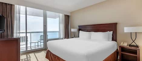 Habitación de hotel con una cama grande, un escritorio, una silla y vistas al océano desde las puertas del balcón.