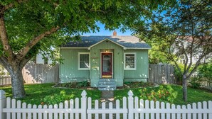 A small, single-story house with a green exterior, a red door, and a white picket fence in front. - Cozy historic downtown CDA home w/ office space. Pet friendly!  (Coeur d'Alene)