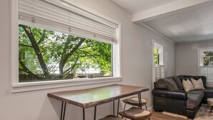 A living room with a wooden table and three bar stools, a dark leather sofa, and a window with blinds overlooking a tree. - Cozy historic downtown CDA home w/ office space. Pet friendly!  (Coeur d'Alene)
