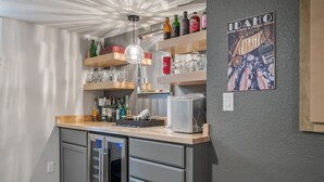 A bar area with shelves holding bottles, glasses, and a coffee machine. There is a refrigerator with a freezer, a wall-mounted light fixture, and a poster on the wall. - Cozy historic downtown CDA home w/ office space. Pet friendly!  (Coeur d'Alene)