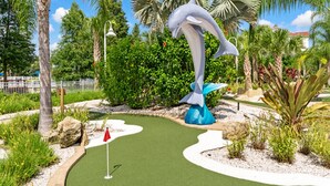 Un campo de minigolf con una gran escultura de delfín, rodeado de plantas tropicales y palmeras.