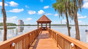 Un muelle de madera que lleva a un pabellón con techo de tejas, rodeado de agua tranquila y árboles.