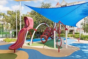 Une aire de jeux avec un toboggan rouge, des structures d’escalade et une voile d’ombrage bleue.