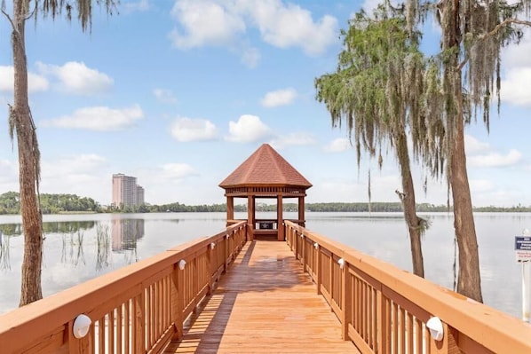 A wooden pier leading to a gazebo by a calm lake with trees and a distant building.