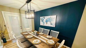 A dining room with a wooden table set for six, white chairs, a world map on the wall, and a hanging light fixture. - Playtime Paradise! Pool, Video Game Room, Pets OK! (Kennewick)