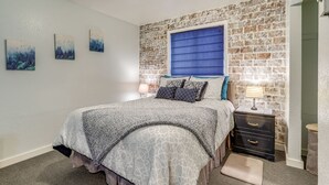 A bedroom with a bed, a nightstand, a lamp, and a window with blue blinds. - Cozy Wasilla Apartment ~ 2 Mi to Downtown! (Wasilla)