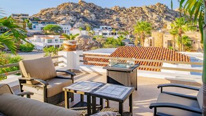 A terrace with outdoor furniture, a view of a rocky mountain, and buildings in the background.