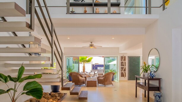 A spacious living area with a high ceiling, a staircase, and a dining area with wicker furniture.
