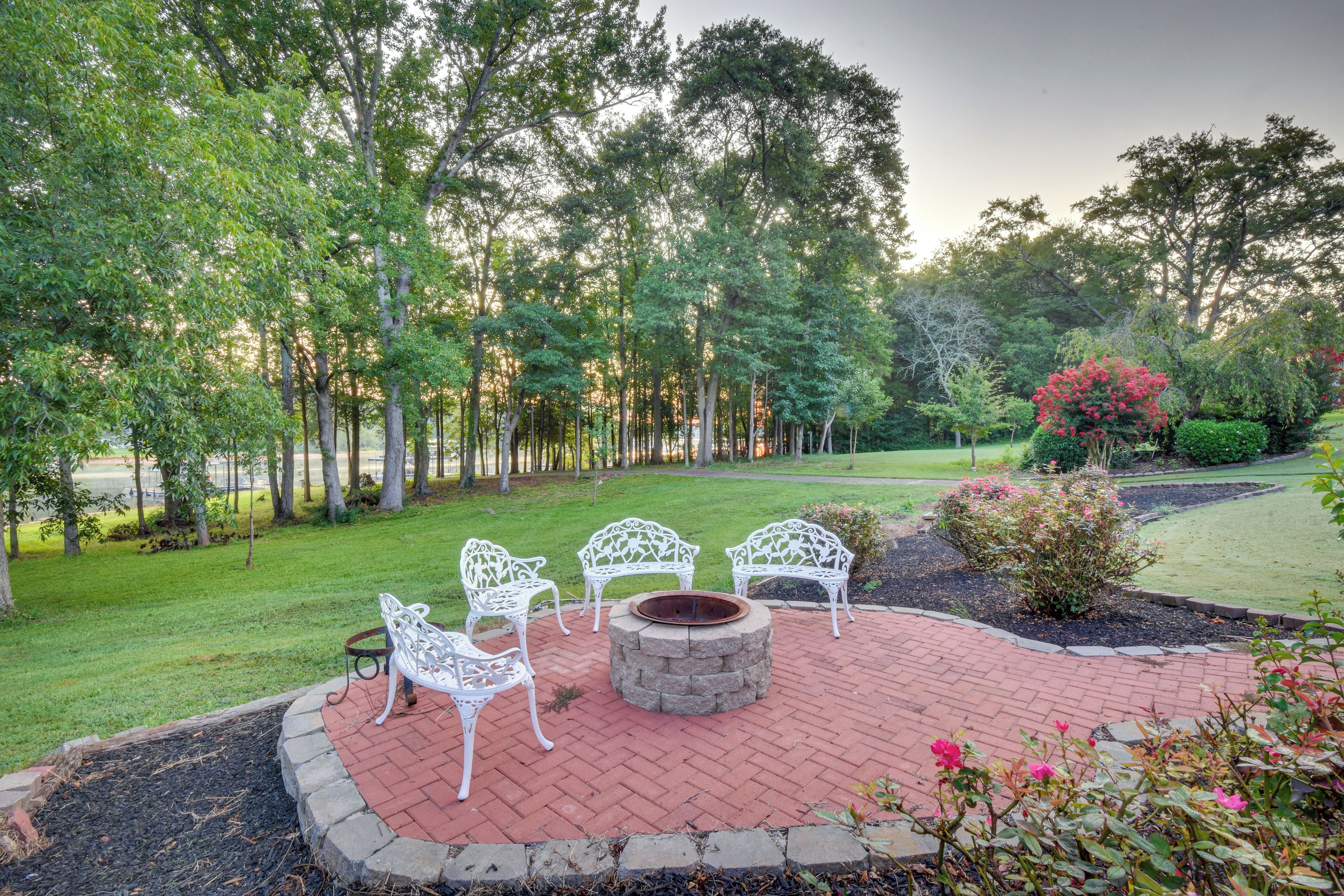 A patio area with white metal chairs and a stone fire pit surrounded by a brick border.