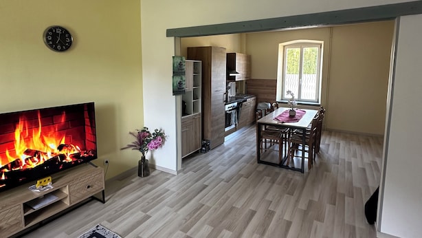 A living room with a flat-screen TV displaying a fireplace, a dining area with a table and chairs, and a kitchen with wooden cabinets and a window. - Can Appart 1 à 8 Pers (Granges-Aumontzey)