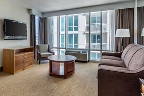 Hotelkamer met een flatscreen televisie, een bank, een stoel, een houten dressoir, een ronde salontafel en grote ramen met uitzicht op de buitenkant van het gebouw.