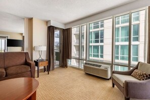 A hotel room with a sofa, a small table with a lamp, a large window with curtains, and a wall-mounted air conditioner.