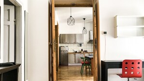 A narrow hallway leading to a kitchen with a wooden door, a red chair, and a bed with a checkered blanket. - Bonerowska 3/48 (Kraków)