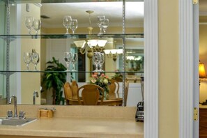 Una cucina con un piano di lavoro in vetro, un lavello e uno specchio che riflette una zona pranzo con sedie e un lampadario.
