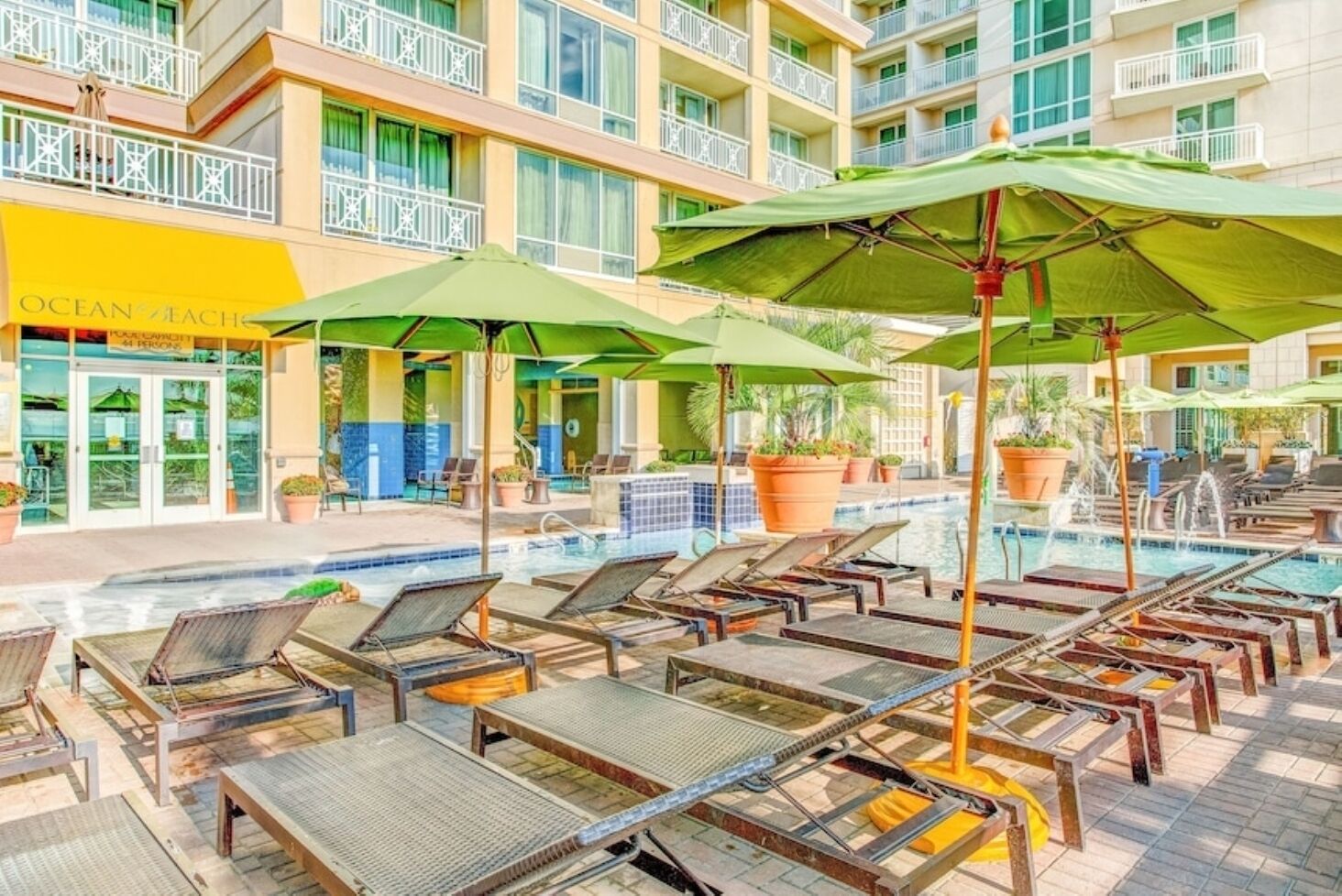 Een zwembadterras met ligbedden en parasols, een gebouw met een bord waarop 'Ocean's Beach' staat, en een betegeld zwembad.