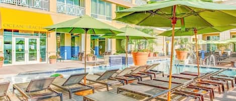 Un área junto a la piscina con sillones de descanso y sombrillas, un edificio con un letrero que dice 'Ocean's Beach', y una piscina con azulejos.