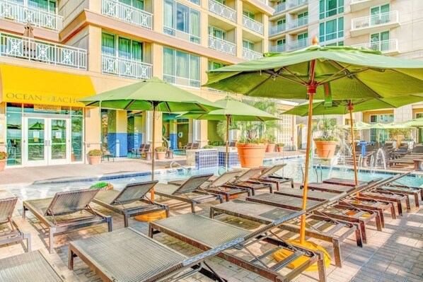 Un área junto a la piscina con sillones de descanso y sombrillas, un edificio con un letrero que dice 'Ocean's Beach', y una piscina con azulejos.