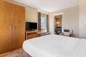 A hotel room with a large bed, a television on a wooden stand, a bathroom with a mirror, and a view of the city through the window. - Diamond Resorts Ocean Beach Club - 3 BDR Suite - Ocean Front (Virginia Beach)