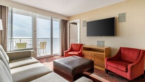 Habitación de hotel con balcón, televisión de pantalla plana, sofá, sillón rojo y cómoda de madera.