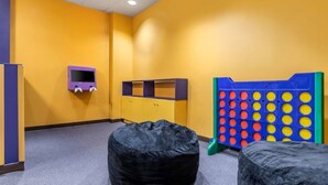 Una habitación con una pared amarilla, un mueble de televisión púrpura, un tablero de juego azul y amarillo, y dos puffs negros.