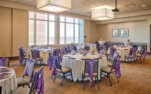 Une salle de banquet avec des tables rondes, des chaises et des rubans violets, dotée de grandes fenêtres et d’un éclairage au plafond.