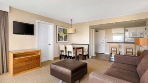 Een hotelkamer met een flatscreen televisie, een houten dressoir, een bruin leren poef, een bruine bank, een eettafel met stoelen en een kitchenette.