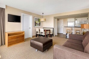 Une chambre d’hôtel équipée d’un téléviseur à écran plat, d’une commode en bois, d’un pouf en cuir marron, d’un canapé marron, d’une table à manger avec des chaises et d’une kitchenette.