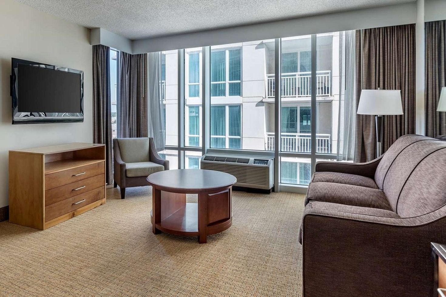 A hotel room with a flat-screen TV, a sofa, a chair, a wooden dresser, a round coffee table, and large windows with a view of the building's exterior.