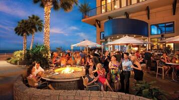 A group of people gathered around a fire pit in an outdoor setting with a building in the background.