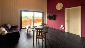 A spacious room with a large sliding glass door leading to a balcony, a wooden ceiling, a purple wall, a round table with chairs, and a clock. - Quietly located apartment on the pond on the upper floor with balcony (Nideggen)