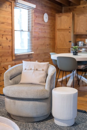 A cozy room with a grey armchair, a white ottoman, and a wooden dining table with chairs. - Red Feather Cabins 250 (Boone)