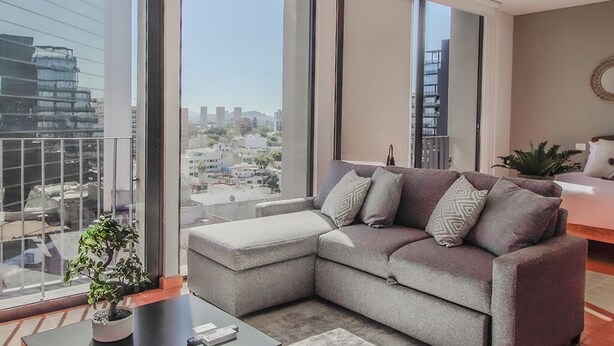 A modern living room with a grey sectional sofa, a small potted plant, and a view of a cityscape through large windows. - Tribu Chapultepec 67 (Guadalajara)